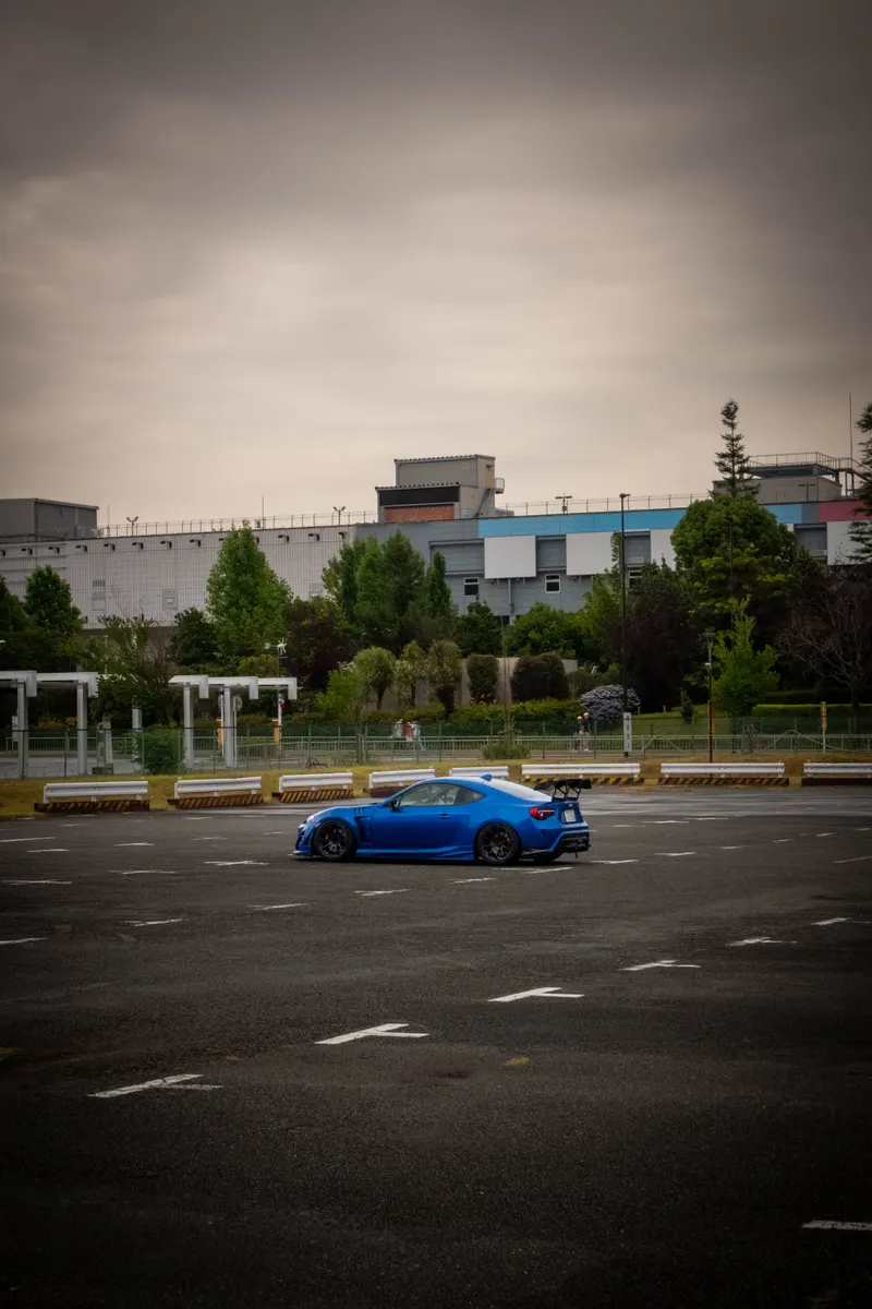 A blue widebody Subaru BRZ parked alone in an empty car park under a heavy grey sky.