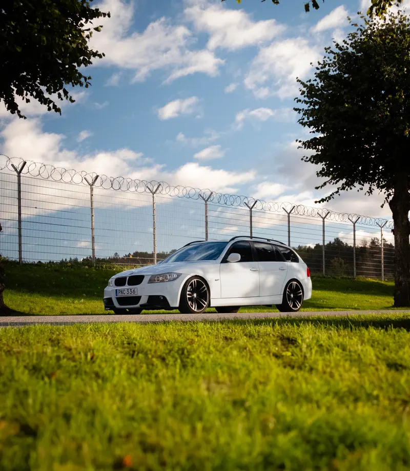 A white BMW 320d Touring on a roadside, shot low through summer grass with a blue sky behind.